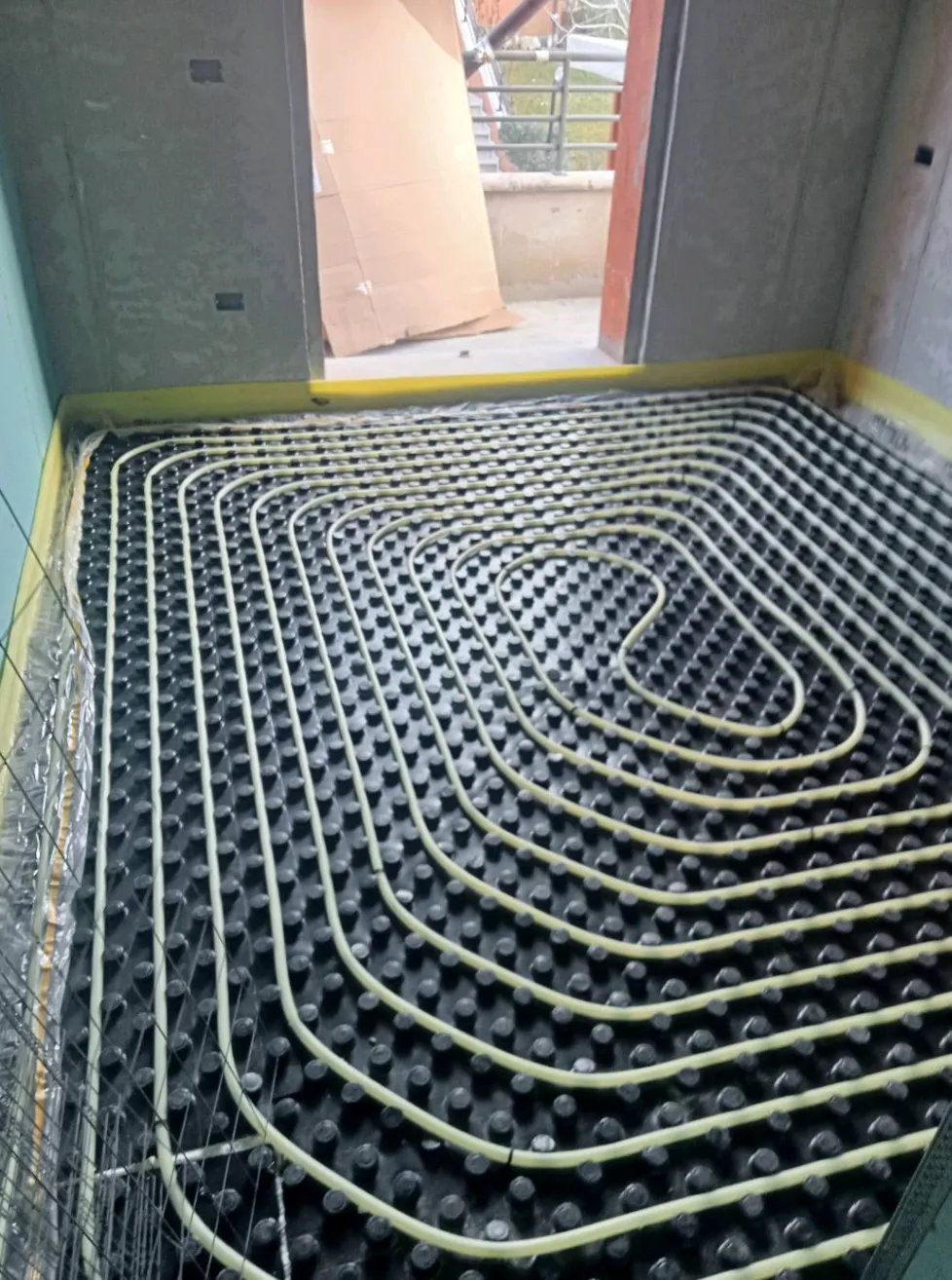Room under construction with installed underfloor heating tubing arranged in a spiral pattern on a black plastic base.