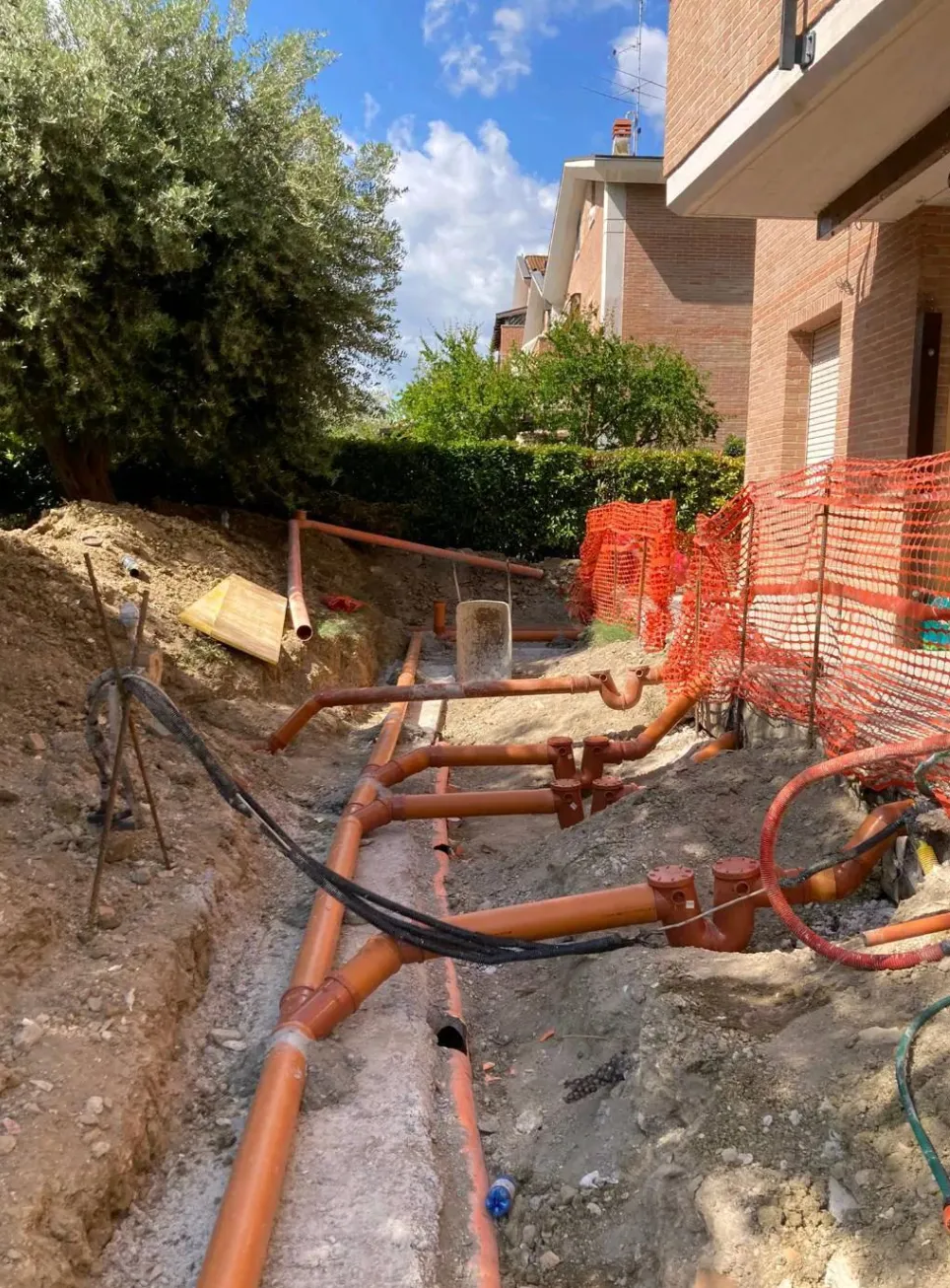 Construction site next to a building with multiple orange underground pipes installed in a trench surrounded by dirt and orange safety fencing.