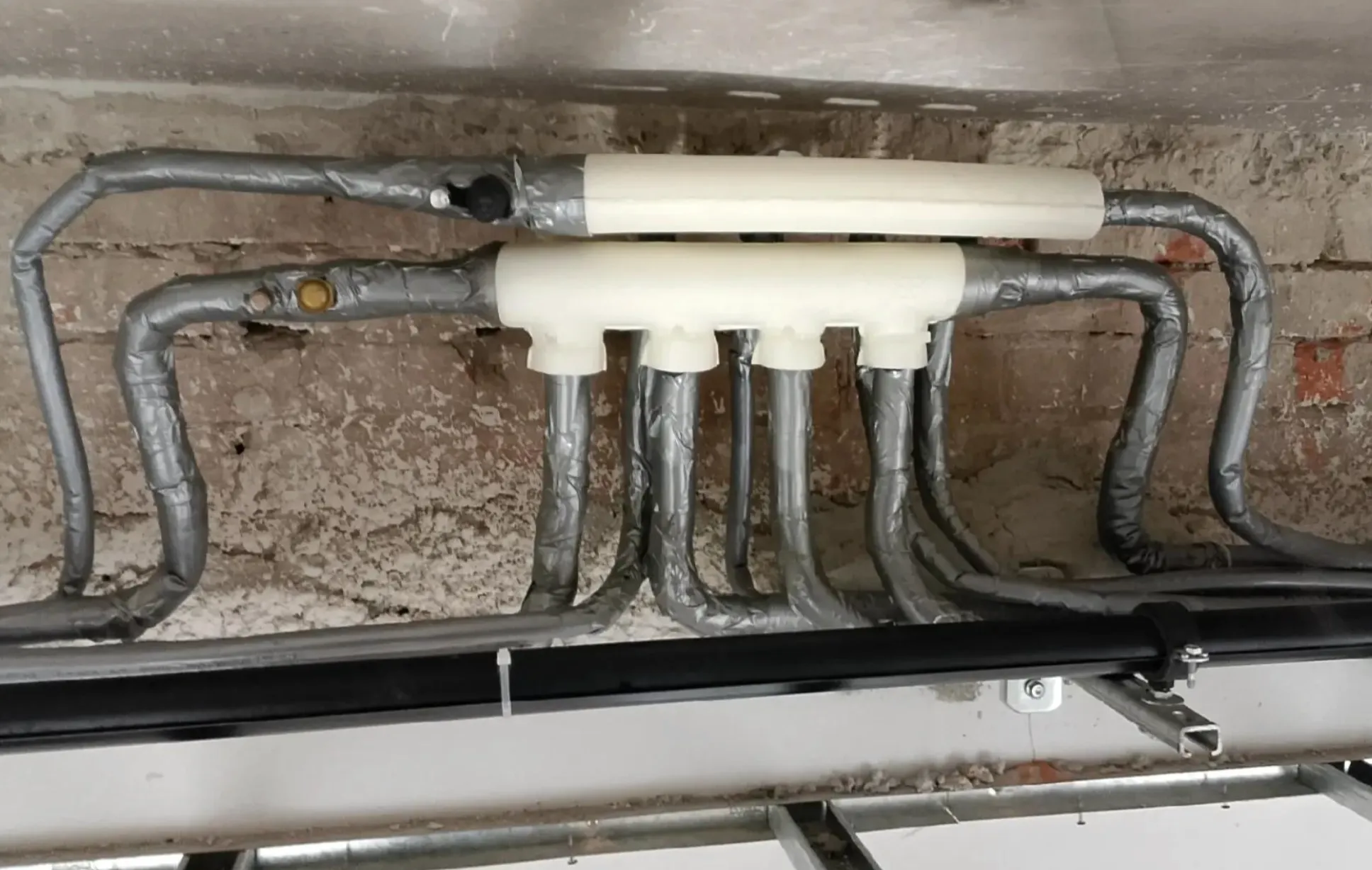 Insulated plumbing pipes connected to a white manifold against a rough brick wall.