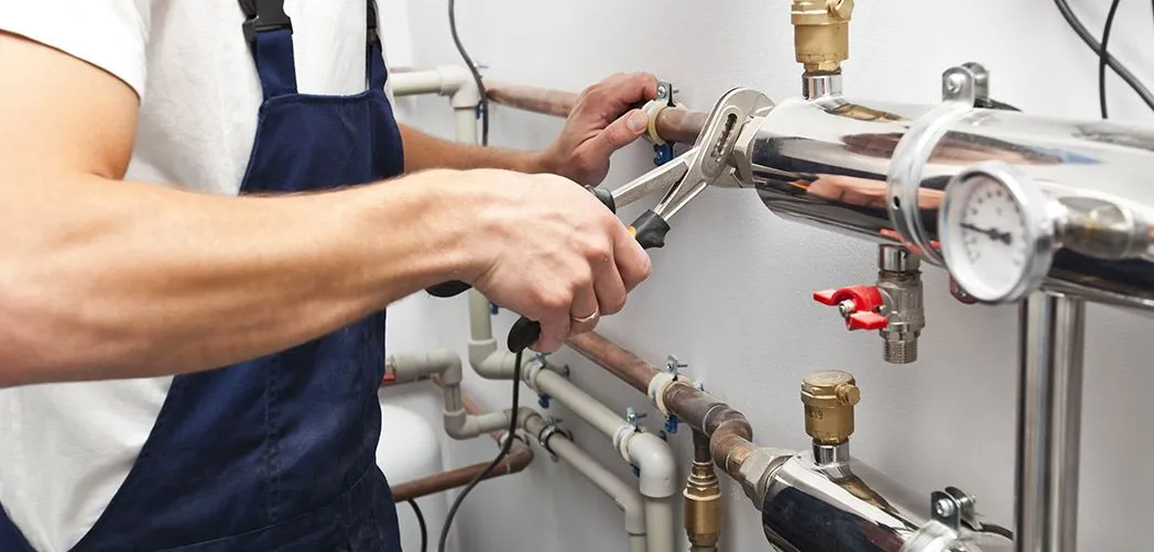 Technician in white shirt and blue overalls tightening a pipe fitting with a wrench on a wall-mounted plumbing system with gauges and valves.